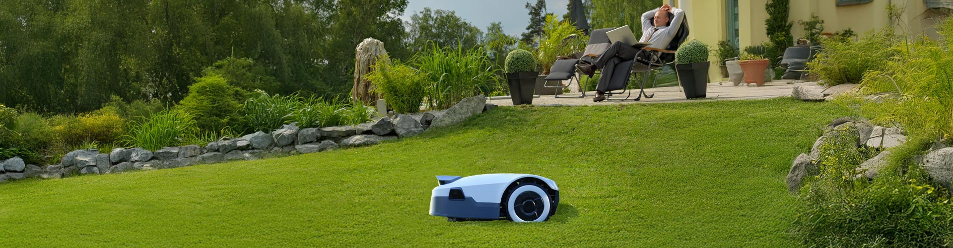 Anthbot Mähroboter mäht den Rasen, während ein entspannter Gartenbesitzer im Hintergrund auf der Terrasse sitzt